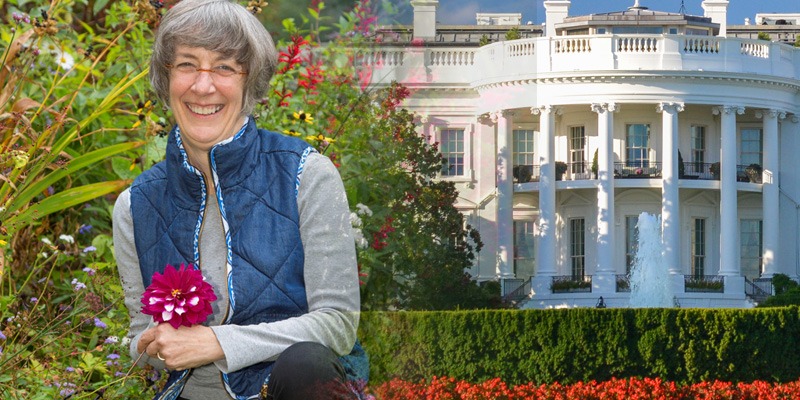 Marta McDowell squatting and holding a flower in front of the White House smiling at the camera